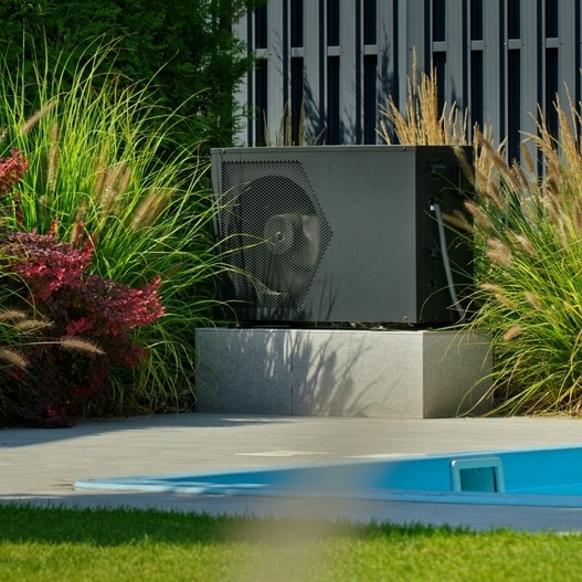 Pompe à chaleur de piscine : chauffage efficace et moderne Pompe à chaleur de piscine noire installée sur un socle dans un jardin paysager moderne près du bassin.